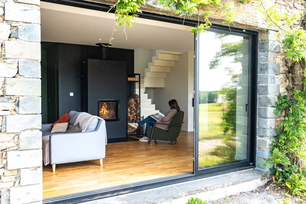 Vue extérieure à travers une baie vitrée d'un poêle de masse scandinave Tulikivi installé dans une maison rénovée en pierre.